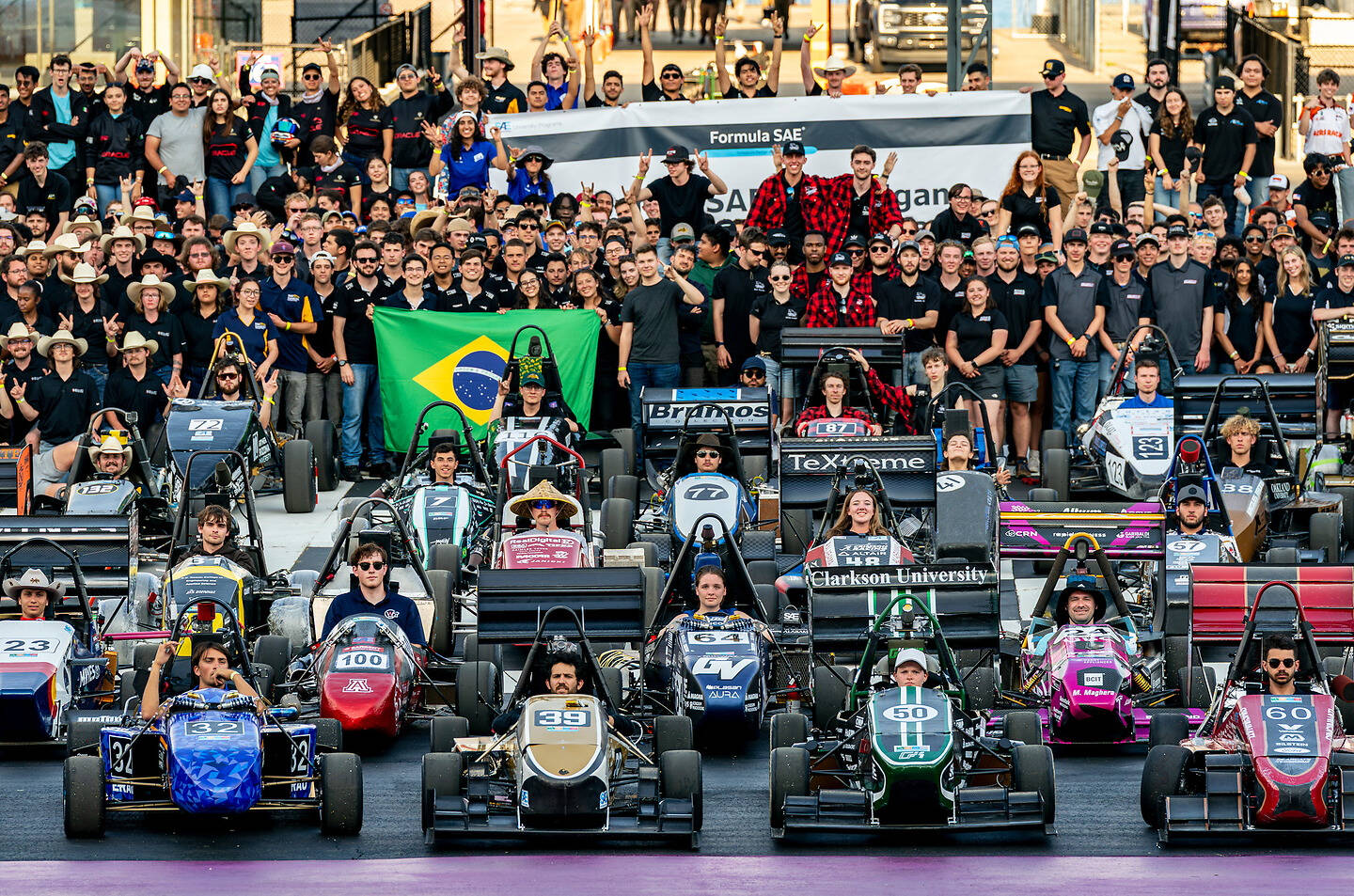 A large group of racing teams and spectators gather at a Formula SAE event, with numerous open-wheel race cars lined up on the track and a Brazilian flag prominently displayed.